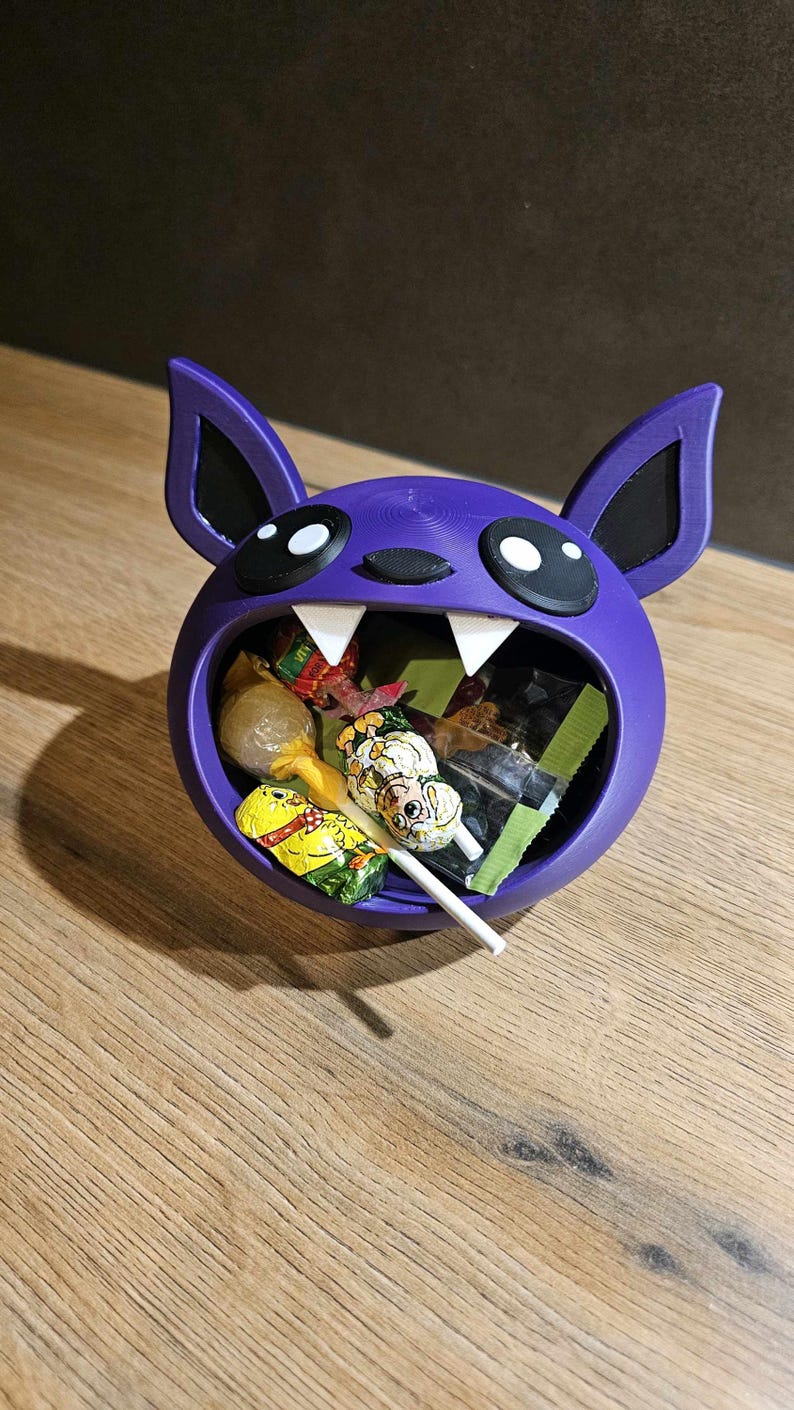 May include: A purple bat-shaped candy bowl filled with various wrapped candies and a lollipop. The bowl has large white fangs, black eyes, and ears. The candies are in colorful wrappers. The bowl sits on a wooden surface.