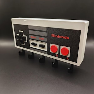 May include: A white and black key holder designed to look like a classic Nintendo controller. The key holder has the Nintendo logo in red, red A and B buttons, and black hooks for keys. It is designed to be mounted on a wall.