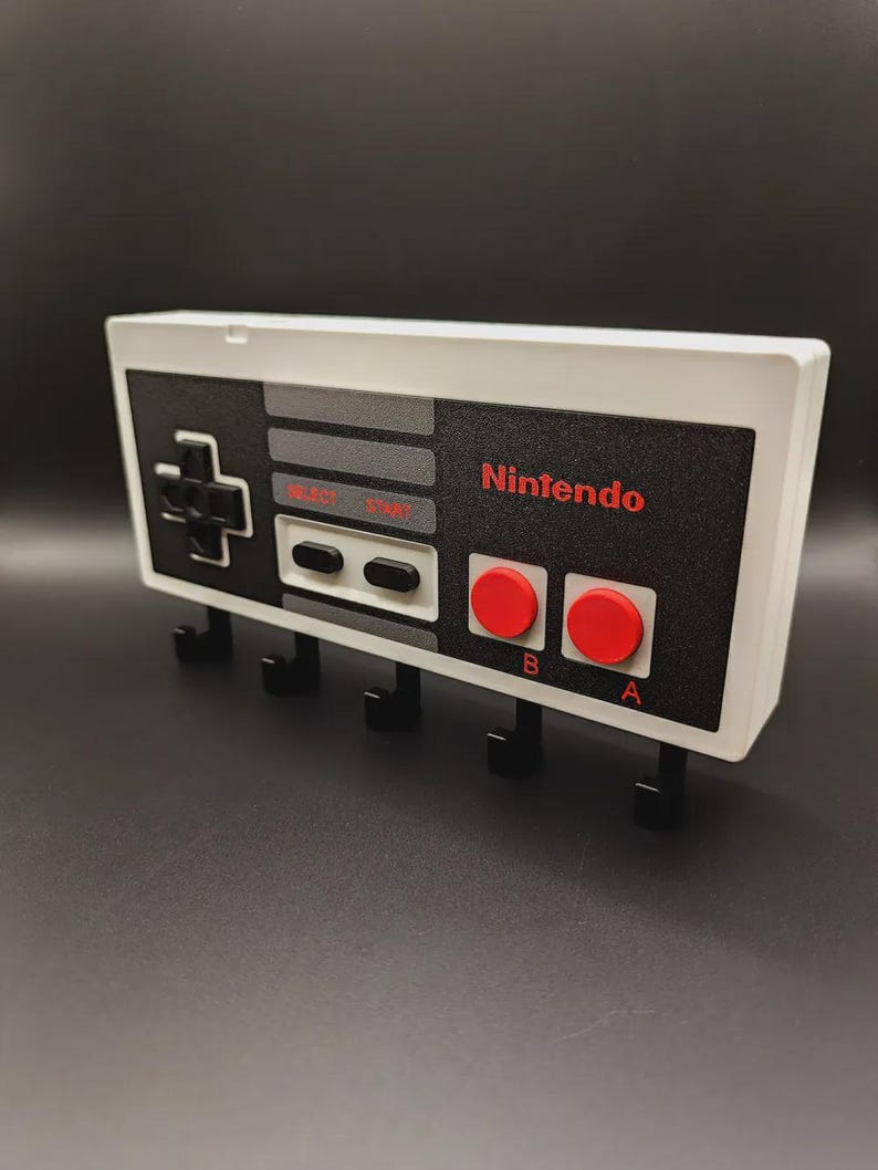 May include: A white and black key holder designed to look like a classic Nintendo controller. The key holder has the Nintendo logo in red, red A and B buttons, and black hooks for keys. It is designed to be mounted on a wall.
