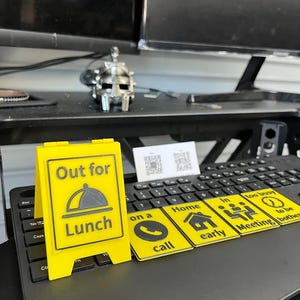 May include: A yellow and black desk sign that reads "Out for Lunch" with a graphic of a plate and a dome. Also, small yellow and black signs with the text "on a call", "Home early", "Meeting", and "too busy to be bothered".