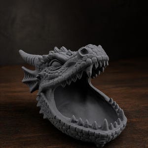 May include: A gray dragon head sculpture with an open mouth, revealing a bowl-shaped interior. The dragon has detailed scales, horns, and sharp teeth. The sculpture is made of a solid material and sits on a wooden surface.
