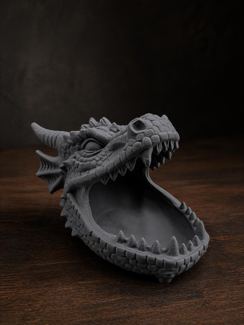 May include: A gray dragon head sculpture with an open mouth, revealing a bowl-shaped interior. The dragon has detailed scales, horns, and sharp teeth. The sculpture is made of a solid material and sits on a wooden surface.