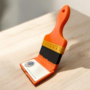 Puede incluir: Porta tarjetas de visita en forma de pincel naranja con un &aacute;rea de cerdas de pincel negra y un borde dorado. El porta tarjetas est&aacute; abierto y se puede ver una tarjeta de visita con el texto "Color Without Compromise".