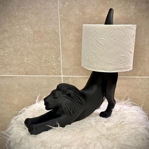 Pode incluir: Um porta-papel higi&eacute;nico preto em forma de le&atilde;o. O le&atilde;o est&aacute; numa pose de alongamento, sustentando um rolo de papel higi&eacute;nico branco. O suporte &eacute; feito de um material s&oacute;lido e repousa sobre uma superf&iacute;cie branca e fofa. O fundo &eacute; uma parede de azulejos de tons neutros.
