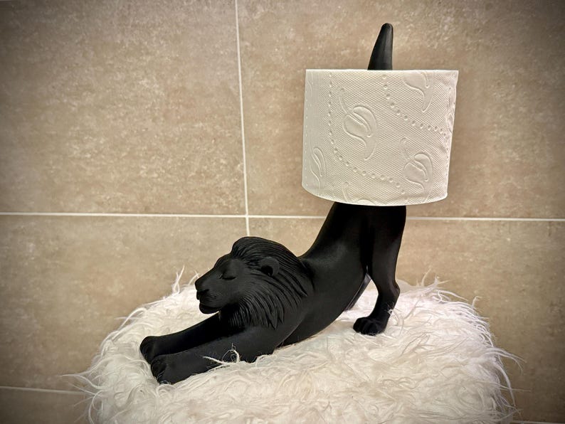 Pode incluir: Um porta-papel higi&eacute;nico preto em forma de le&atilde;o. O le&atilde;o est&aacute; numa pose de alongamento, sustentando um rolo de papel higi&eacute;nico branco. O suporte &eacute; feito de um material s&oacute;lido e repousa sobre uma superf&iacute;cie branca e fofa. O fundo &eacute; uma parede de azulejos de tons neutros.