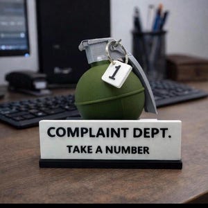 Könnte beinhalten: Ein neuartiges Schreibtischschild mit der Aufschrift "COMPLAINT DEPT. TAKE A NUMBER" mit einer grünen Granate und einem Nummer-Eins-Schild. Das Schild ist weiß mit schwarzem Text, und die Granate ist olivgrün.