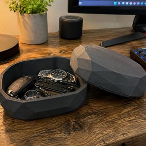 Puede incluir: Una bandeja de valet de color gris oscuro, de forma geom&eacute;trica, con una tapa a juego, abierta para revelar un reloj, llaves, una multiherramienta y un anillo. La bandeja est&aacute; sobre un escritorio de madera, junto a un teclado y un monitor.