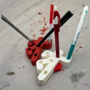 May include: A white and red 3D printed pen holder shaped like a stick figure. The holder has multiple slots for pens, pencils, and other writing utensils. A black pen, a red pen, a blue pen, and a gray screwdriver are in the holder.