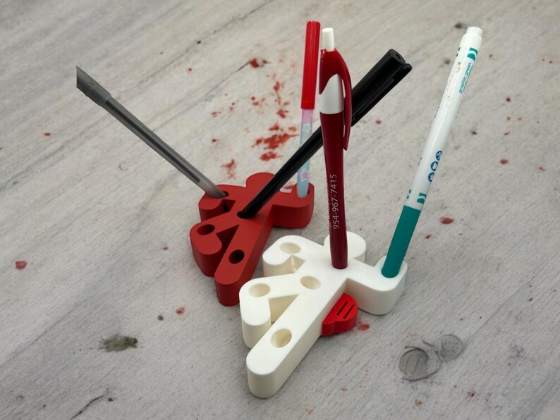 May include: A white and red 3D printed pen holder shaped like a stick figure. The holder has multiple slots for pens, pencils, and other writing utensils. A black pen, a red pen, a blue pen, and a gray screwdriver are in the holder.