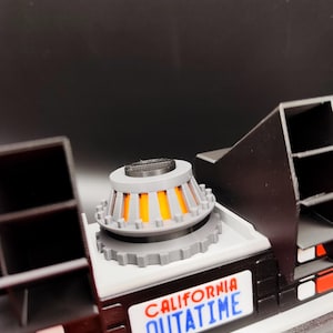 Pode incluir: Modelo detalhado de uma m&aacute;quina do tempo DeLorean, com um capacitor de fluxo cinza e laranja, carroceria preta e placa branca com a inscri&ccedil;&atilde;o "CALIFORNIA OUTATIME".