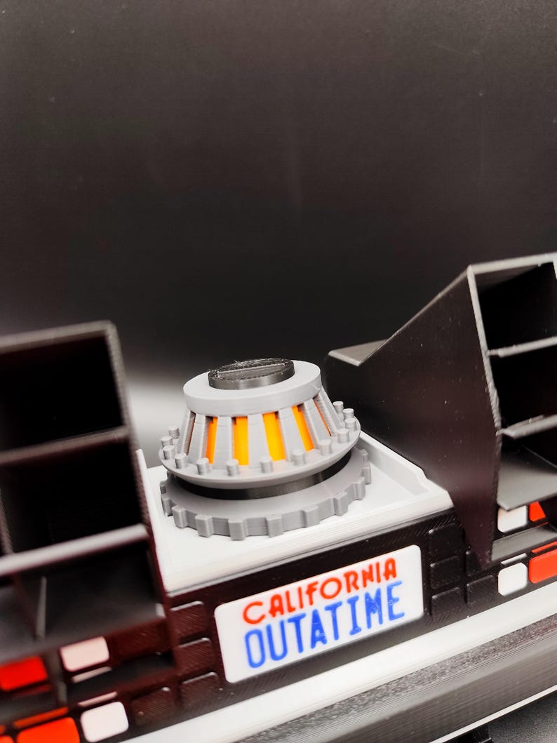 Pode incluir: Modelo detalhado de uma m&aacute;quina do tempo DeLorean, com um capacitor de fluxo cinza e laranja, carroceria preta e placa branca com a inscri&ccedil;&atilde;o "CALIFORNIA OUTATIME".