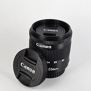 May include: A black Canon camera lens with a lens cap. The lens has a silver band with the text "Canon" and "55mm" printed on it. The lens cap also has the word "Canon" printed on it.