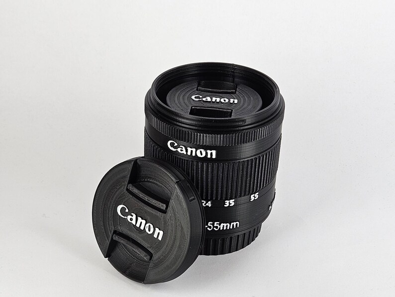 May include: A black Canon camera lens with a lens cap. The lens has a silver band with the text "Canon" and "55mm" printed on it. The lens cap also has the word "Canon" printed on it.