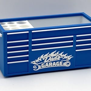 May include: A blue toolbox with white accents and the text "Dad's Garage" in a flame design. The toolbox has a handle and a section with circular compartments. The design includes horizontal white stripes.