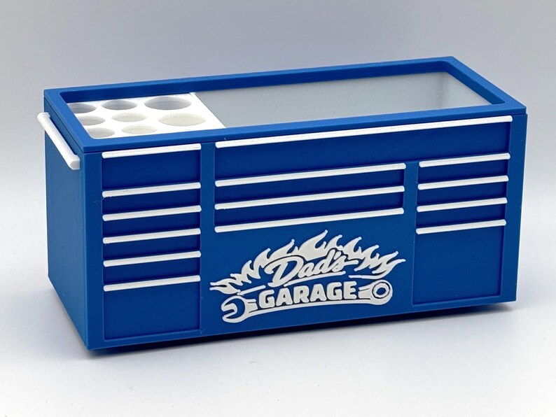 May include: A blue toolbox with white accents and the text "Dad's Garage" in a flame design. The toolbox has a handle and a section with circular compartments. The design includes horizontal white stripes.