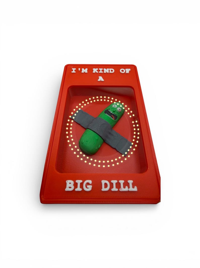 May include: A red display case with the text "I'M KIND OF A" and "BIG DILL" in white. Inside, a green pickle character with a gray strap is surrounded by a circle of lights.