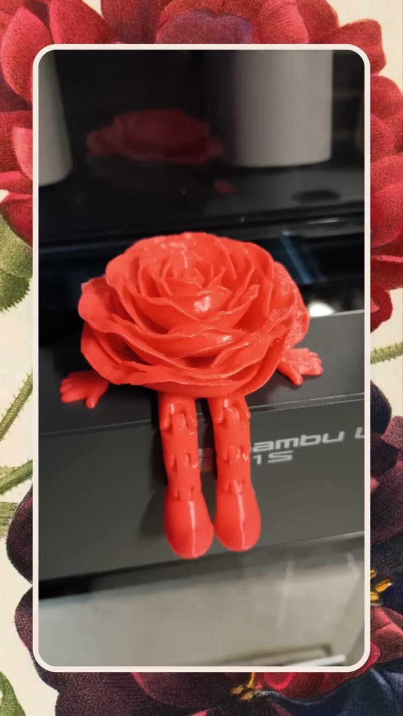 May include: A 3D printed red rose with legs. The rose is sitting on a black surface with the word "ambu" printed on it.