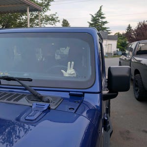 Puede incluir: Un Jeep Wrangler azul con un guante blanco haciendo el signo de la paz a trav&eacute;s del parabrisas. El veh&iacute;culo est&aacute; estacionado afuera en un d&iacute;a nublado. Se ve un espejo lateral negro.