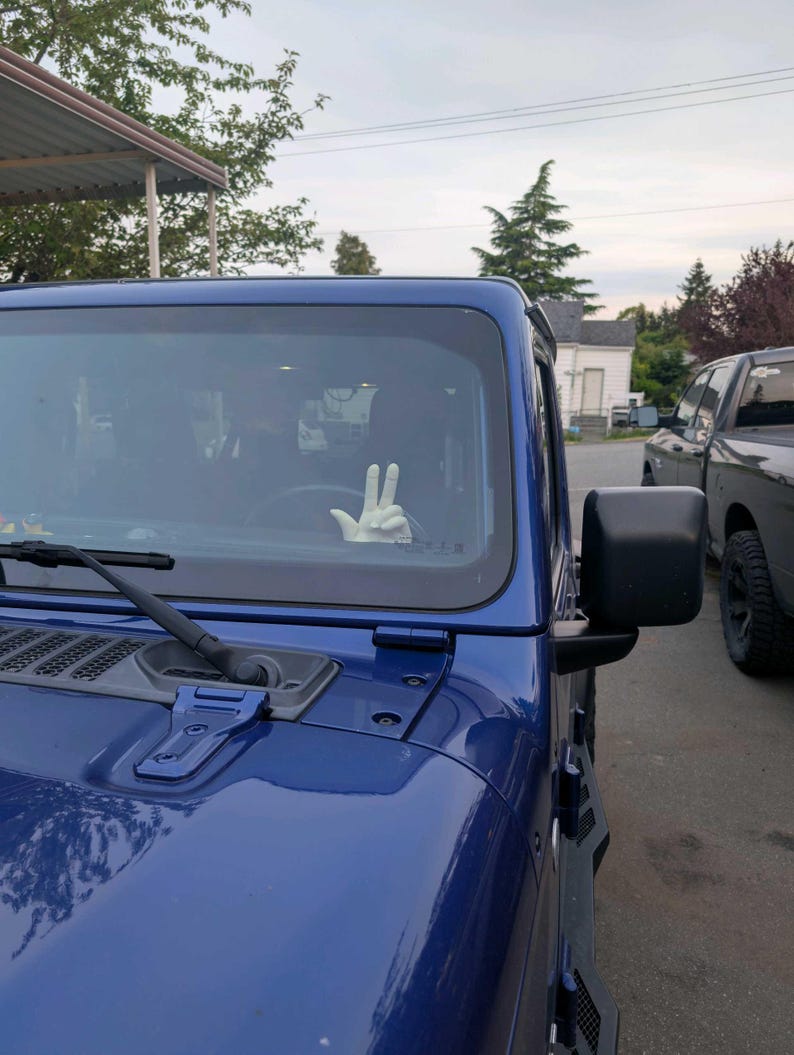 Puede incluir: Un Jeep Wrangler azul con un guante blanco haciendo el signo de la paz a trav&eacute;s del parabrisas. El veh&iacute;culo est&aacute; estacionado afuera en un d&iacute;a nublado. Se ve un espejo lateral negro.