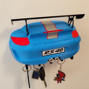 K&ouml;nnte beinhalten: Blauer, an der Wand befestigter Schl&uuml;sselhalter in Form eines Autos. Er hat einen schwarzen Spoiler und einen silbernen Streifen. Auf dem Nummernschild steht "RS 4.0". Mehrere Schl&uuml;ssel und Schl&uuml;sselanh&auml;nger h&auml;ngen unten.