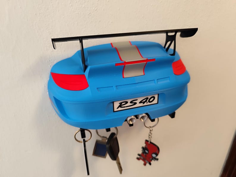 K&ouml;nnte beinhalten: Blauer, an der Wand befestigter Schl&uuml;sselhalter in Form eines Autos. Er hat einen schwarzen Spoiler und einen silbernen Streifen. Auf dem Nummernschild steht "RS 4.0". Mehrere Schl&uuml;ssel und Schl&uuml;sselanh&auml;nger h&auml;ngen unten.