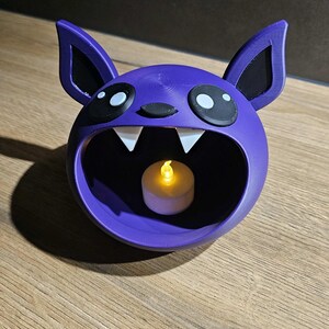 May include: A purple bat-shaped candle holder with large eyes, ears, and fangs. The interior holds a small, lit, battery-operated candle. The holder is made of a 3D-printed material and sits on a wooden surface.