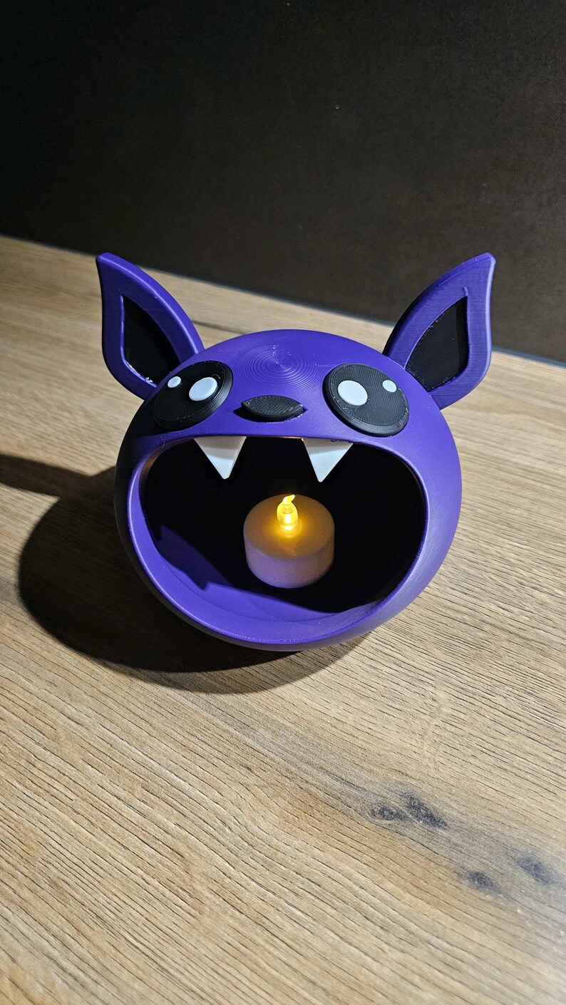 May include: A purple bat-shaped candle holder with large eyes, ears, and fangs. The interior holds a small, lit, battery-operated candle. The holder is made of a 3D-printed material and sits on a wooden surface.