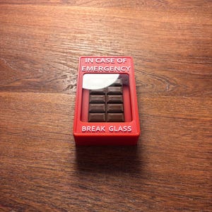 May include: A red plastic box with the text "IN CASE OF EMERGENCY" and "BREAK GLASS" on it. The box contains a chocolate bar.