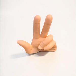 Puede incluir: Escultura de mano de cer&aacute;mica color melocot&oacute;n, mostrando el signo de la paz con los dedos &iacute;ndice y medio levantados. La escultura est&aacute; sobre un fondo blanco liso, enfatizando el gesto de la mano.