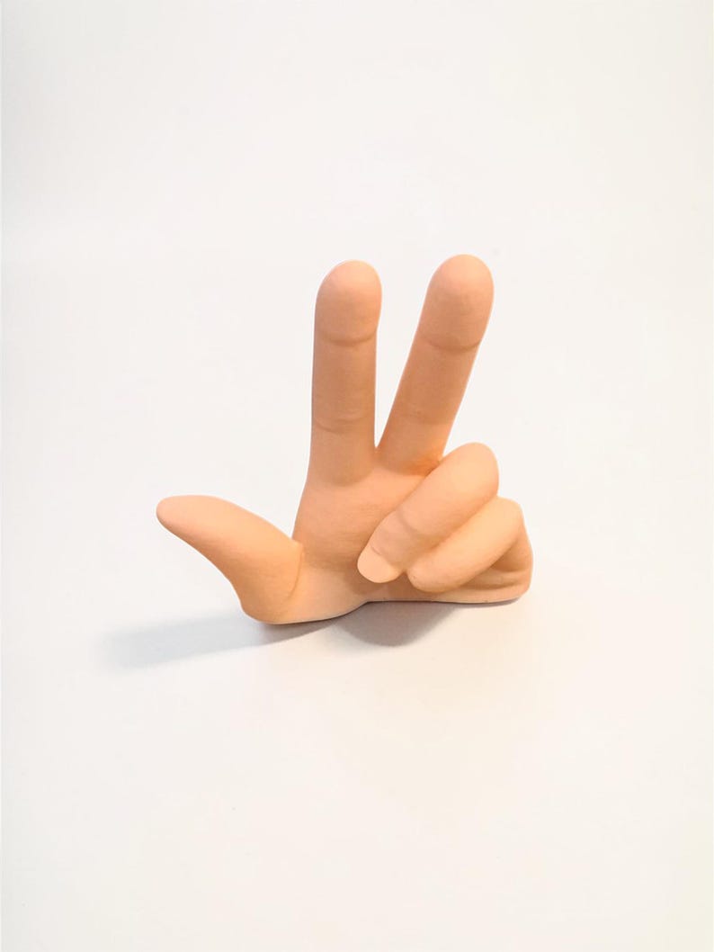 Puede incluir: Escultura de mano de cer&aacute;mica color melocot&oacute;n, mostrando el signo de la paz con los dedos &iacute;ndice y medio levantados. La escultura est&aacute; sobre un fondo blanco liso, enfatizando el gesto de la mano.