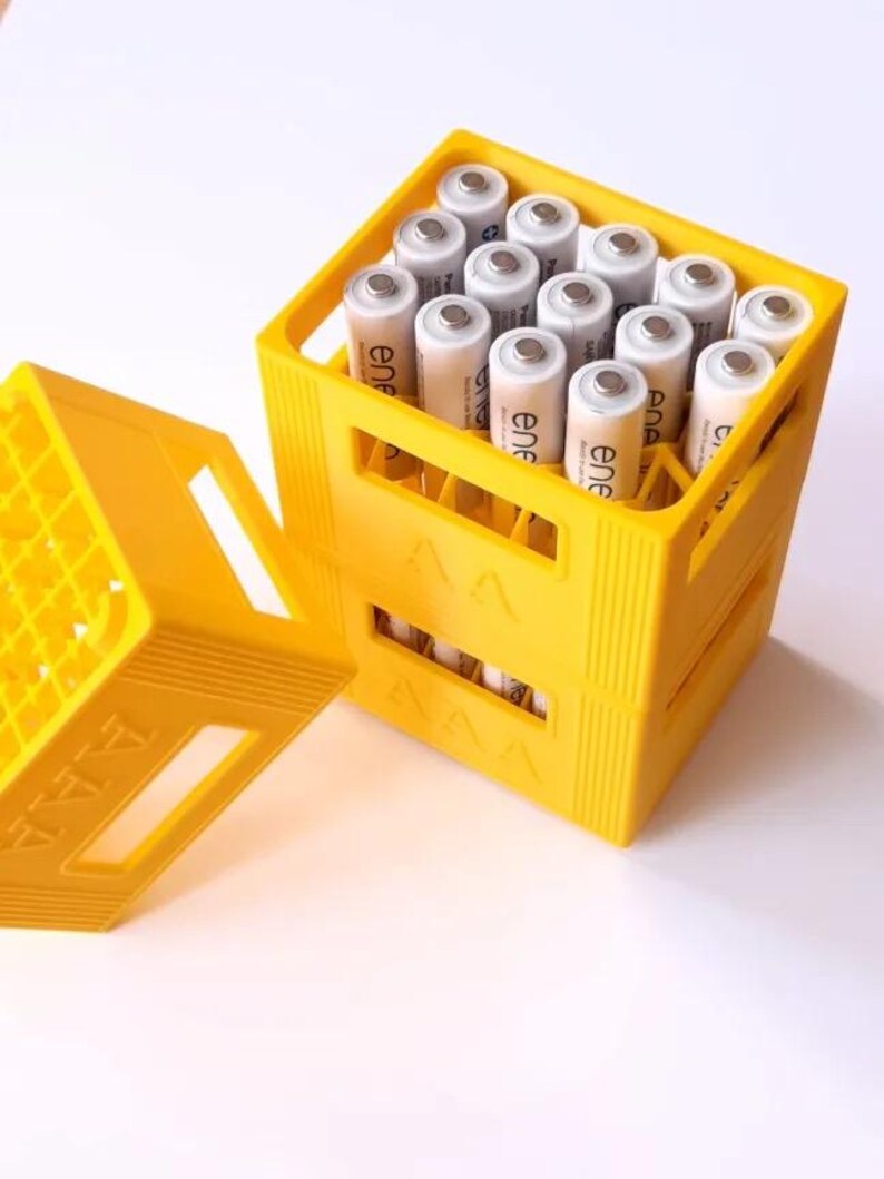 May include: A yellow plastic container filled with AAA batteries. The container is open, revealing the batteries inside. The batteries are cylindrical with silver tops and white bodies. The container is on a white surface.