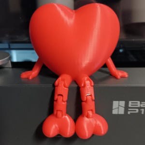 Puede incluir: Un coraz&oacute;n rojo impreso en 3D con piernas articuladas sentado en una superficie gris. La superficie tiene el texto "Bambu P1S" impreso en ella.