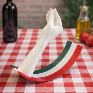 Op de afbeelding: Een witte handgebaarsculptuur met een basis in de kleuren van de Italiaanse vlag, geplaatst op een rood-wit geblokt tafelkleed. Een fles rode wijn, olijfolie en tomaten staan op de achtergrond. Een decoratief item.