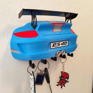 K&ouml;nnte beinhalten: Ein blauer, auto-f&ouml;rmiger Schl&uuml;sselhalter mit einem schwarzen Spoiler und Schl&uuml;sselhaken. Der Schl&uuml;sselhalter hat rote und silberne Akzente, und der Text "RS 4.0" ist sichtbar. Mehrere Schl&uuml;ssel und Schl&uuml;sselanh&auml;nger h&auml;ngen an den Haken.
