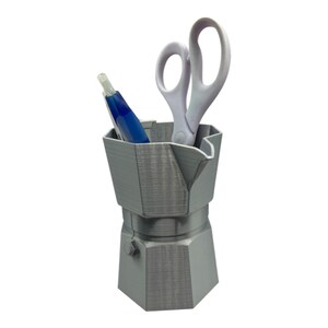 May include: A gray, 3D-printed desk organizer shaped like a stovetop espresso maker. It holds a blue pen and a pair of white scissors. The organizer has a hexagonal base and a spout-like feature on top.