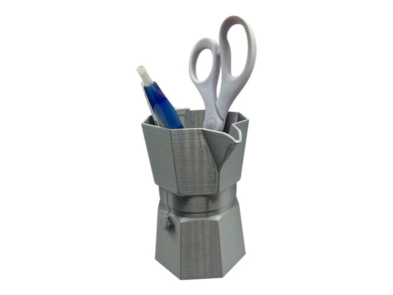 May include: A gray, 3D-printed desk organizer shaped like a stovetop espresso maker. It holds a blue pen and a pair of white scissors. The organizer has a hexagonal base and a spout-like feature on top.