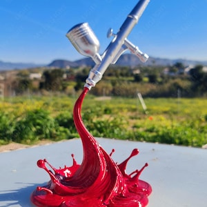 May include: A silver airbrush spraying a vibrant red liquid, creating a splash effect. The image is set against a backdrop of a clear blue sky and a blurred landscape.