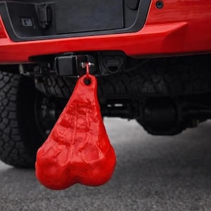 May include: A bright red, textured, anatomical-shaped novelty accessory hanging from the trailer hitch receiver of a red pickup truck. The object features a rounded, dual-lobed design with a small circular attachment point at the top for mounting.