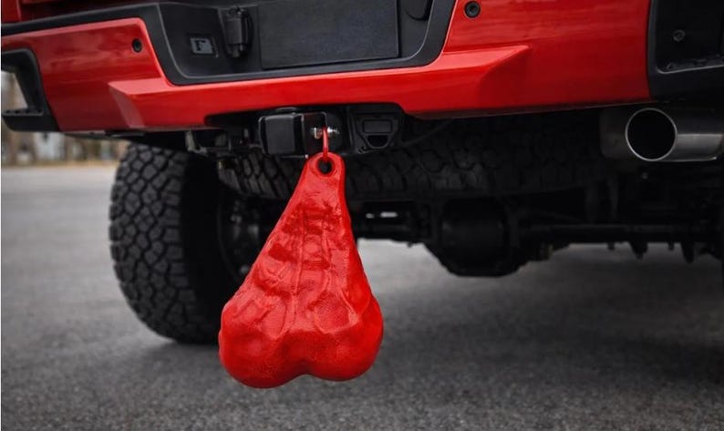 May include: A bright red, textured, anatomical-shaped novelty accessory hanging from the trailer hitch receiver of a red pickup truck. The object features a rounded, dual-lobed design with a small circular attachment point at the top for mounting.