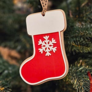 May include: A Christmas stocking ornament with a red body, white cuff, and a white snowflake design. The ornament is made of wood and has a brown ribbon for hanging. The background is a blurred Christmas tree.