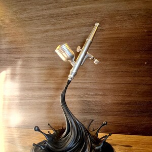 May include: A silver airbrush with a black paint splash base. The airbrush is detailed with a cup, nozzle, and trigger. The base is a stylized splash of black paint, with droplets extending outwards. The background is a wooden panel.