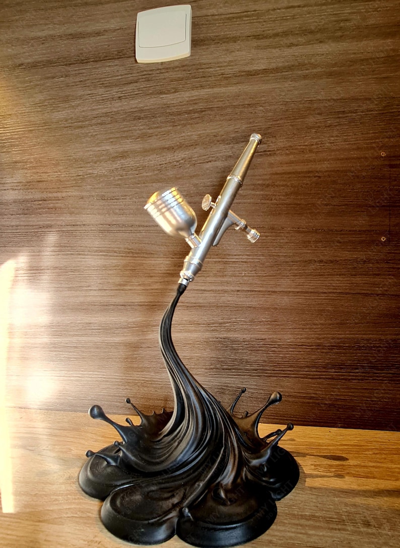 May include: A silver airbrush with a black paint splash base. The airbrush is detailed with a cup, nozzle, and trigger. The base is a stylized splash of black paint, with droplets extending outwards. The background is a wooden panel.