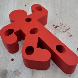 May include: A red, plastic, human-shaped figure with multiple holes. The figure is lying on a light wood surface.