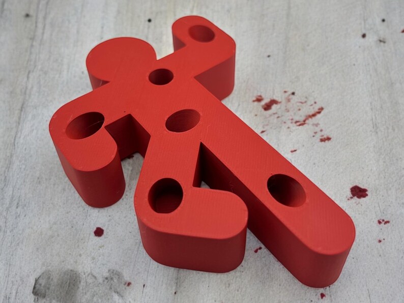 May include: A red, plastic, human-shaped figure with multiple holes. The figure is lying on a light wood surface.