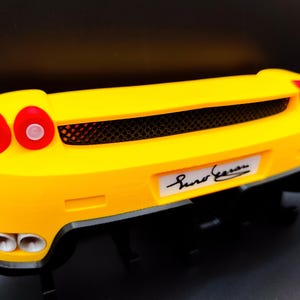 Puede incluir: Parte trasera de un coche modelo amarillo con luces traseras rojas y una parrilla negra. El coche tiene bajos negros y una placa blanca con una firma. Es una r&eacute;plica en miniatura.