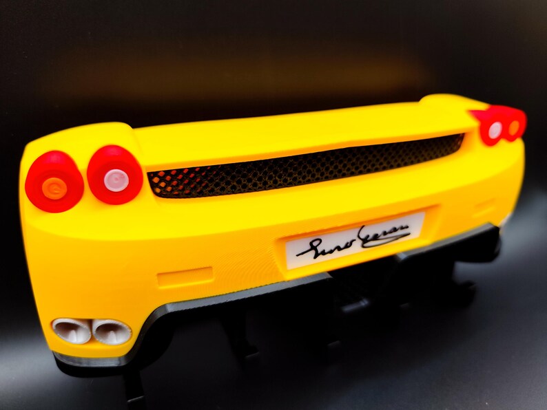 Puede incluir: Parte trasera de un coche modelo amarillo con luces traseras rojas y una parrilla negra. El coche tiene bajos negros y una placa blanca con una firma. Es una r&eacute;plica en miniatura.