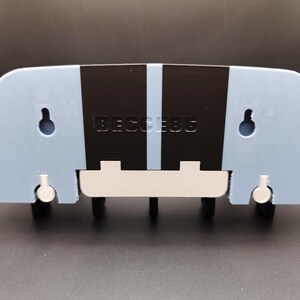 K&ouml;nnte beinhalten: Hellblauer, wandmontierter Schl&uuml;sselhalter mit schwarzen vertikalen Streifen und dem Text "BECESS6". Der Schl&uuml;sselhalter hat eine wei&szlig;e Basis und vier schwarze Schl&uuml;sselhaken. Zwei L&ouml;cher sind zur Montage sichtbar.