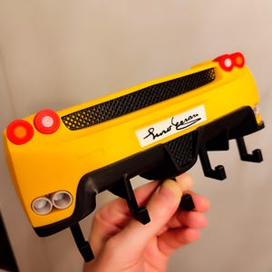Puede incluir: Un gancho de pared amarillo y negro con forma de coche con el texto "Sino Ferrari". El gancho tiene dos luces circulares rojas a cada lado y dos tubos de escape. El gancho est&aacute; sostenido por una mano.
