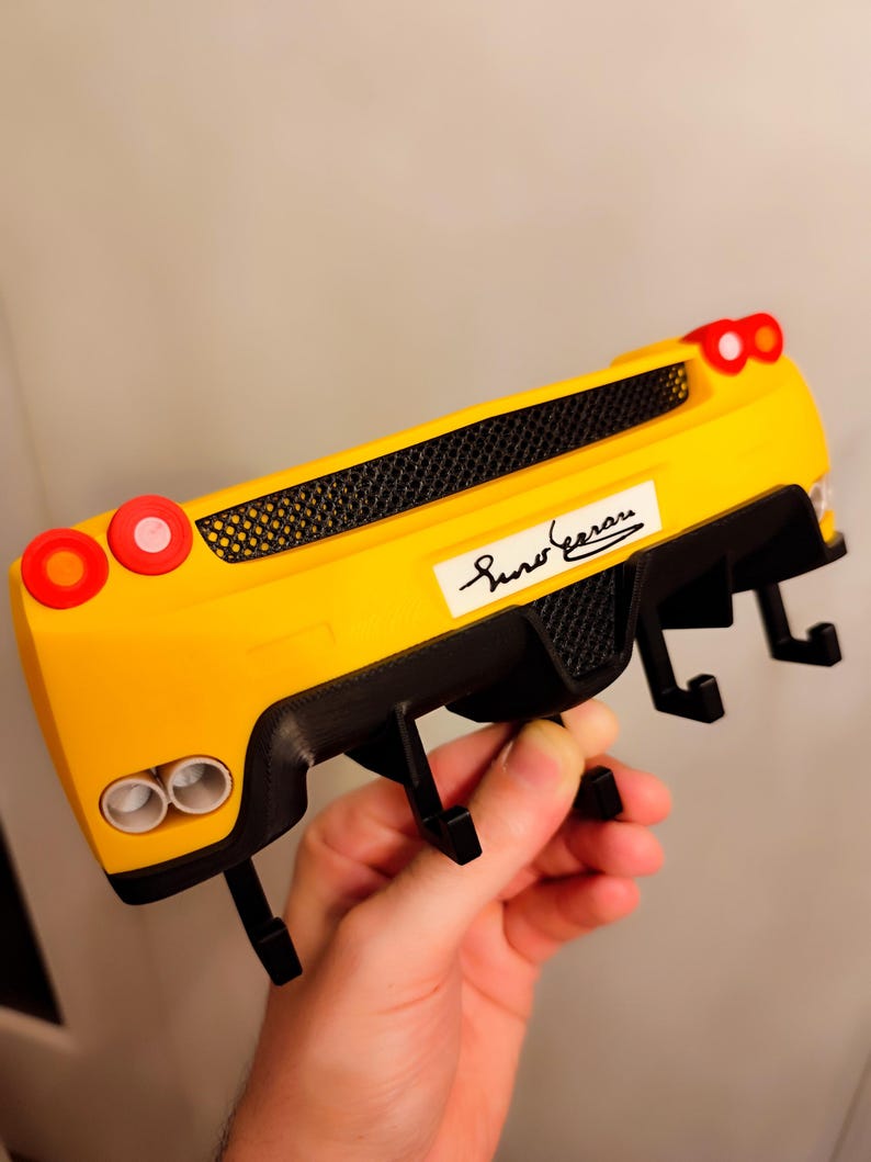 Puede incluir: Un gancho de pared amarillo y negro con forma de coche con el texto "Sino Ferrari". El gancho tiene dos luces circulares rojas a cada lado y dos tubos de escape. El gancho est&aacute; sostenido por una mano.