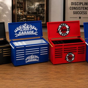 May include: Four miniature toolboxes in black, blue, and red, each with a hinged lid. The black toolbox has "Dad, World's Best Father" text. The blue toolboxes have "Dad's Garage" and "Super Dad" text. The red toolbox has "Best Dad Ever" text.